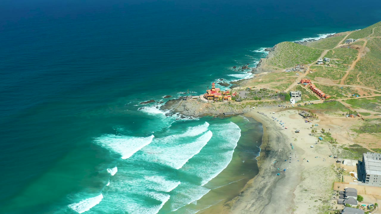 Aerial view at Cerritos Beach with foam bands slightly farther along the shore, deep blue water offshore, small coastal buildings, hillside paths, and no visible people.