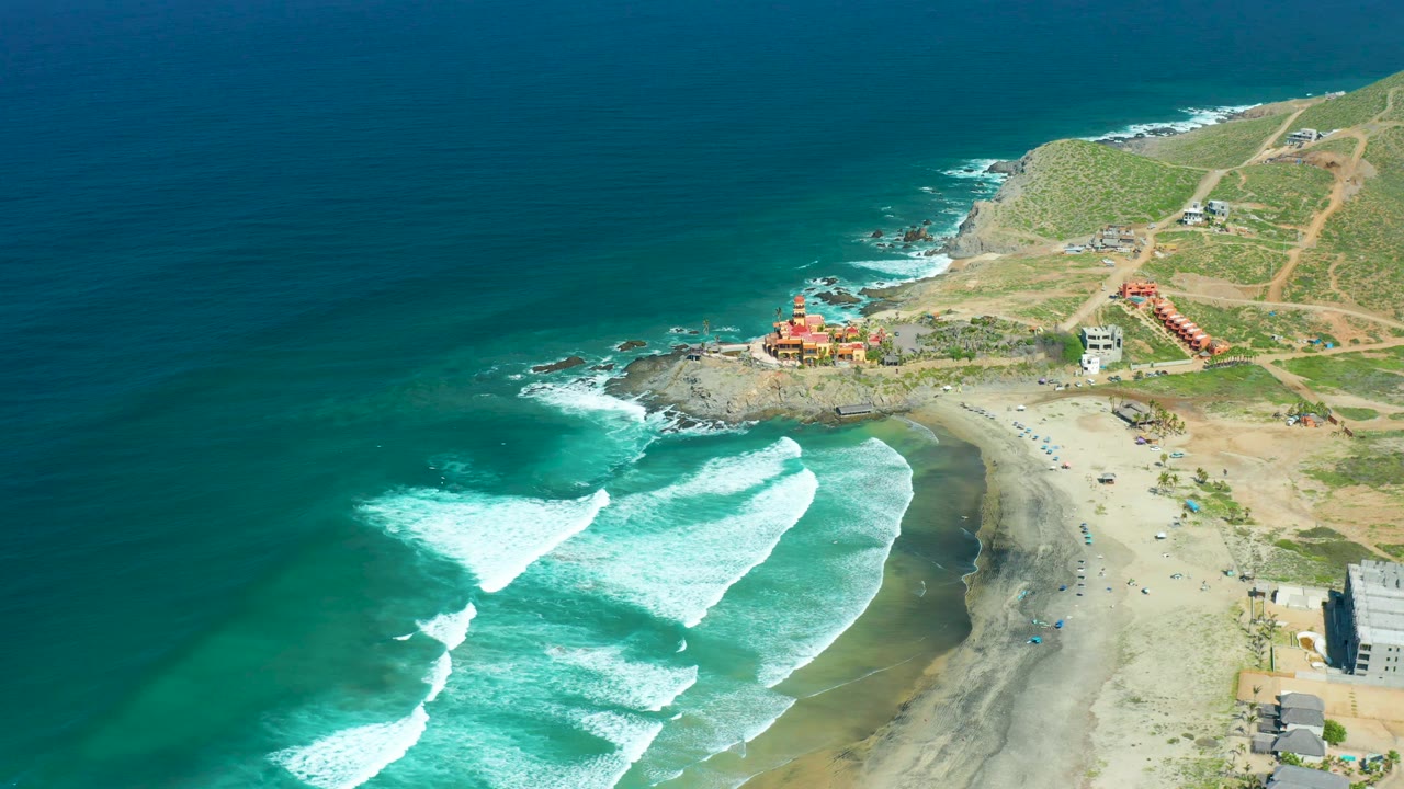 Foamy surf curves toward the beach below coastal hills and roads.