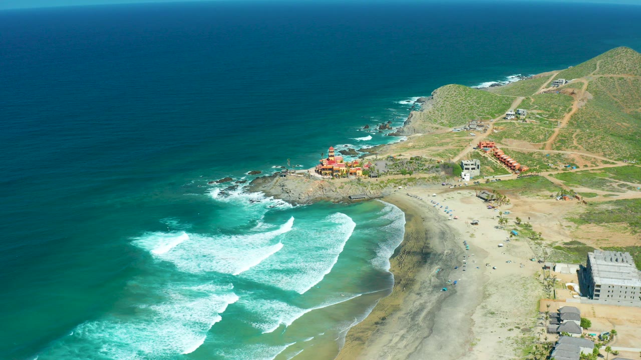 Aerial view of Cerritos Beach in Baja California Sur with turquoise water, white surf, a sandy beach, hillside dirt tracks, small buildings, and no visible people.