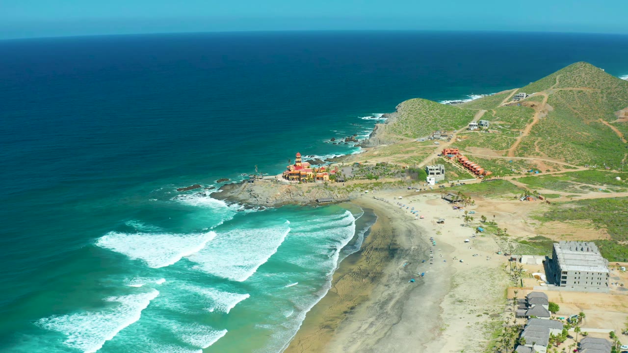 Aerial frame of Cerritos Beach with a bright white wave band pushing closer to the sand, blue-green water filling the left side, buildings near the beach, and no visible people.