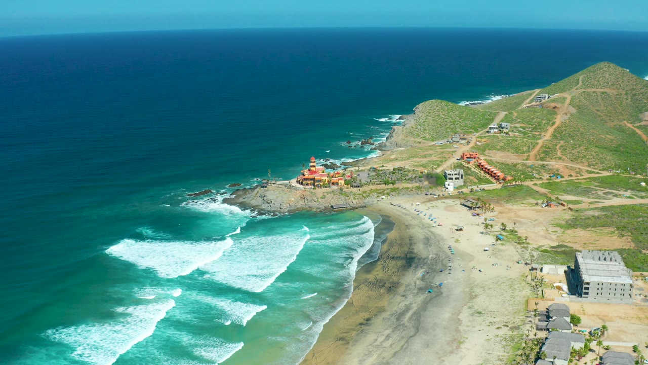 Aerial view at Cerritos Beach showing multiple curved wave lines on the left, a pale sandy beach below, hillside dirt roads at upper right, scattered coastal buildings, and no visible people.