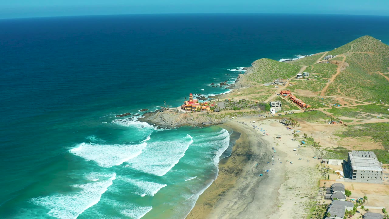 Mid-clip aerial frame over Cerritos Beach with foamy surf spread wider across the lower left, beach and dark wet sand at center, green hills and dirt paths beyond, and no visible people.