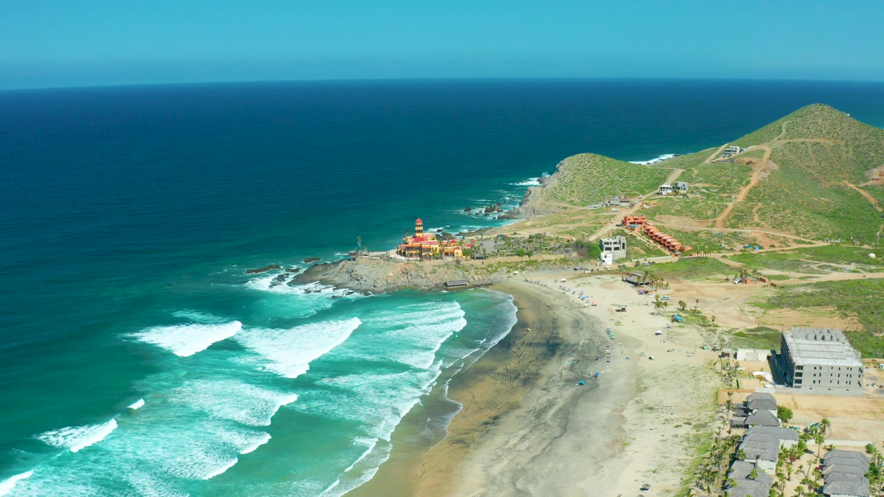 Aerial frame over Cerritos Beach, Baja California Sur, with turquoise water and white surf on the left, sandy shore and hillside buildings to the right, and no visible people.