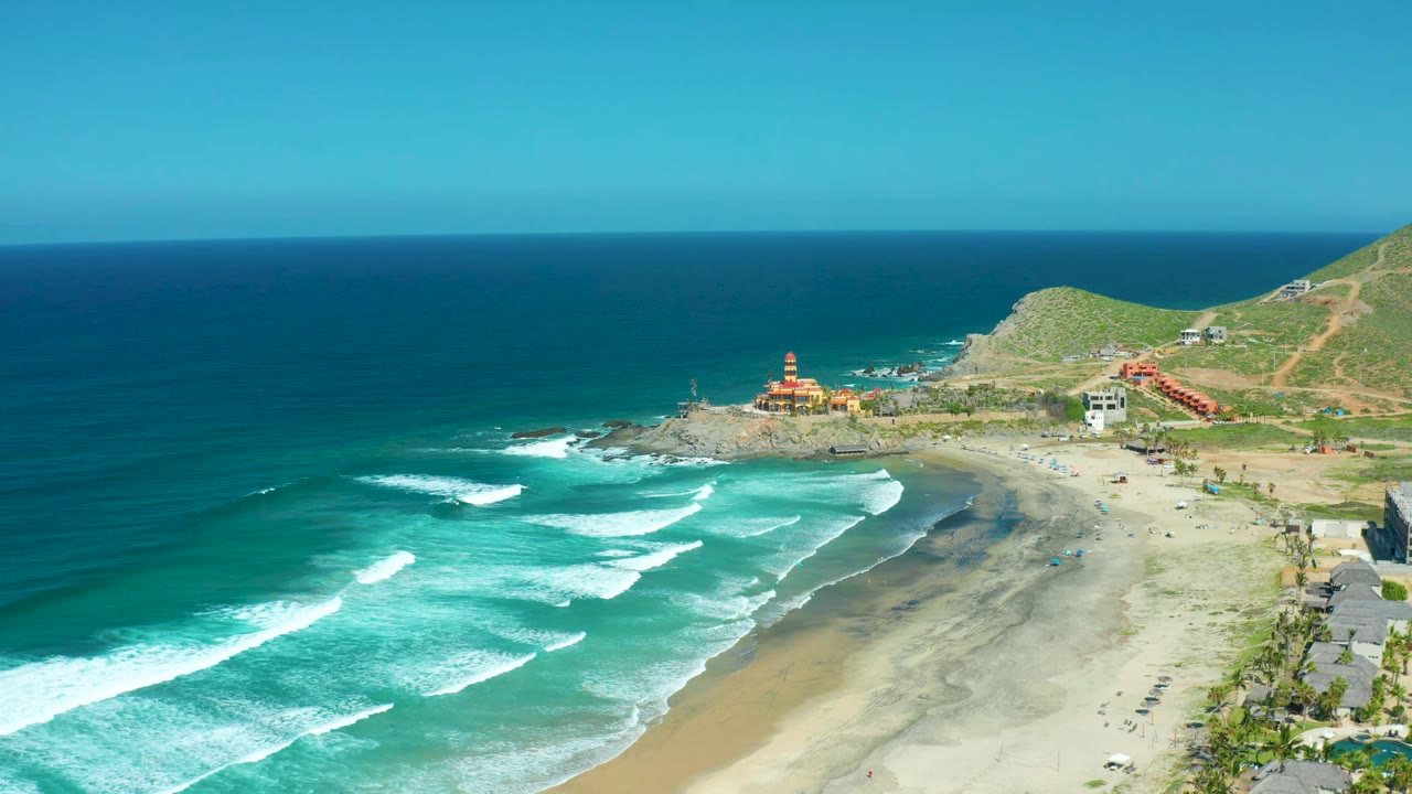 Ocean waves approach a curved sandy beach below a green hillside.