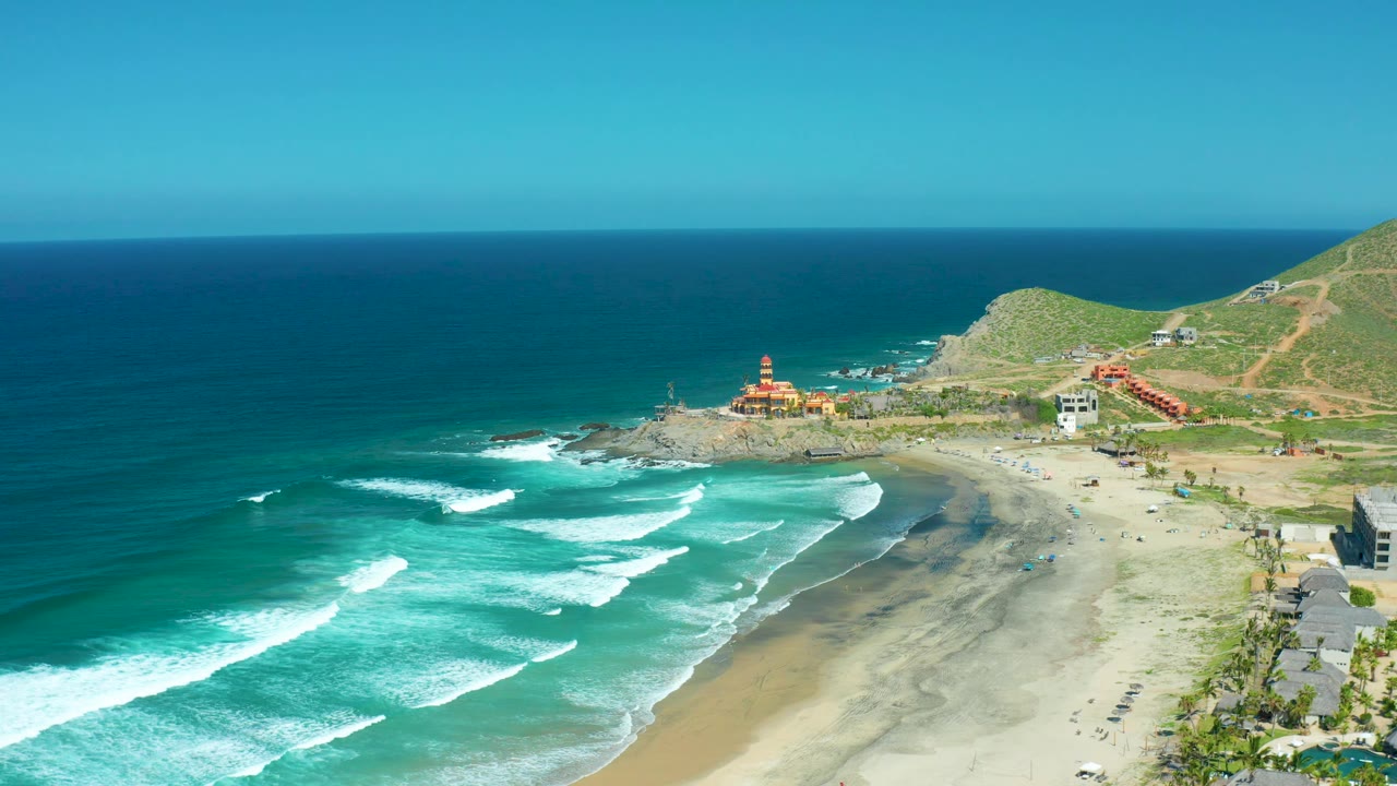 Cerritos Beach in Baja California Sur from above, with surf breaking in staggered white lines, beach sand at right, coastal buildings near the hill, and no visible people.