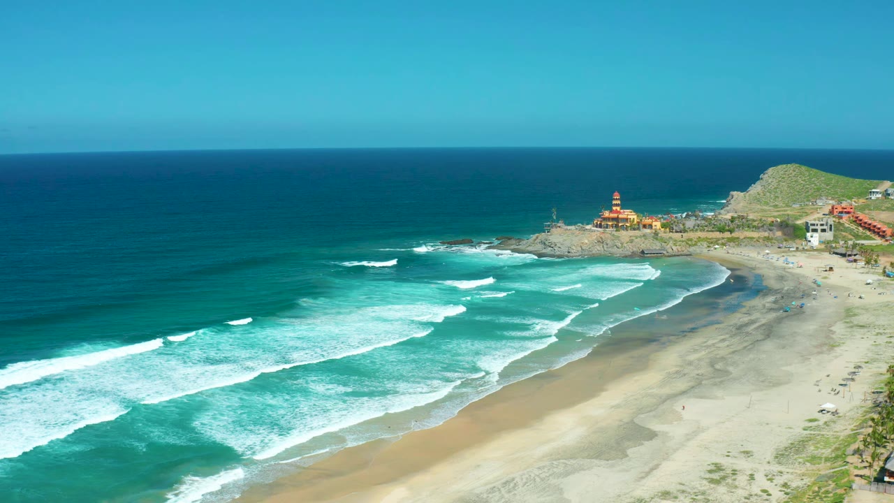 Aerial view one second into the clip at Cerritos Beach, with white surf bands advancing toward wet sand, scattered coastal structures near the hillside, a high ocean horizon, and no visible people.