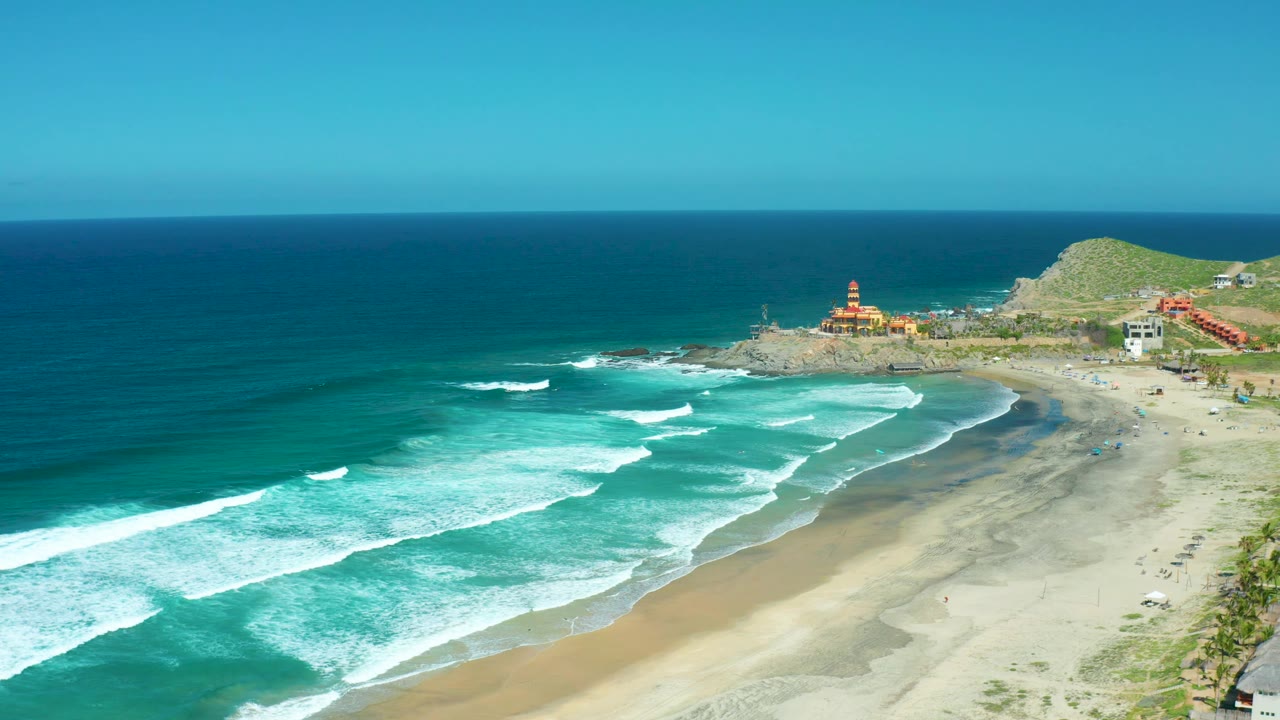 Aerial frame at Cerritos Beach showing a wider sweep of ocean on the left, diagonal whitewater moving toward the beach, a rocky point and low buildings near the hill, and no visible people.