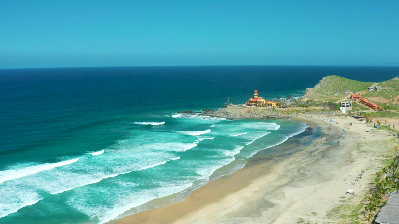 Mid-clip aerial view of Cerritos Beach with foamy waves filling the left foreground, pale sand curving along the right, distant coastal buildings near a green hill, and no visible people.