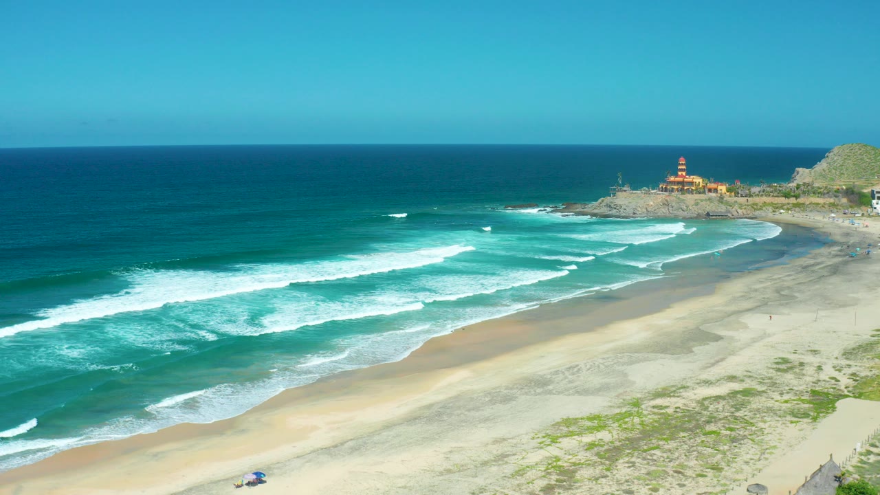 Aerial view of waves breaking on a sandy beach with a lighthouse on a rocky point.