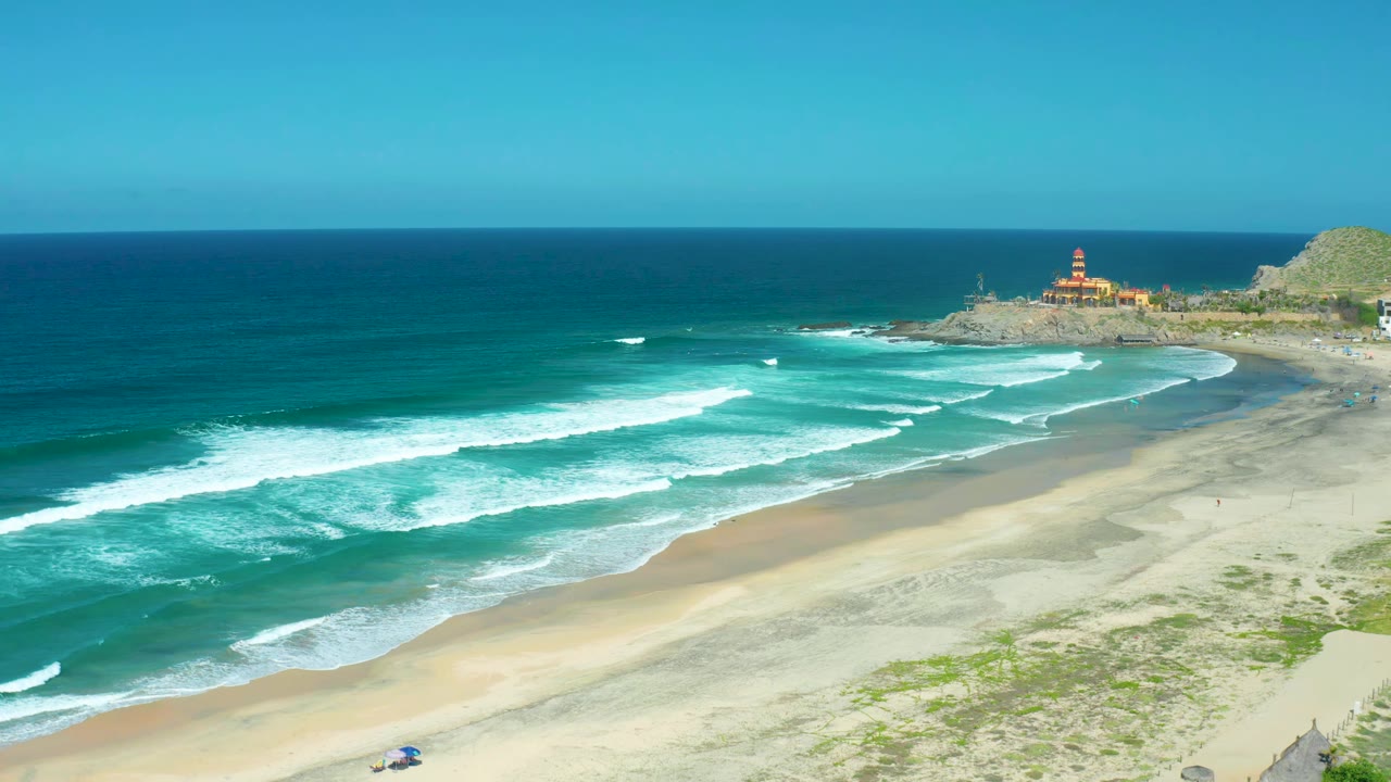 Wide aerial frame over Cerritos Beach shows the surf advancing in parallel white lines, pale sand in the foreground, the lighthouse on the point, and no visible people.