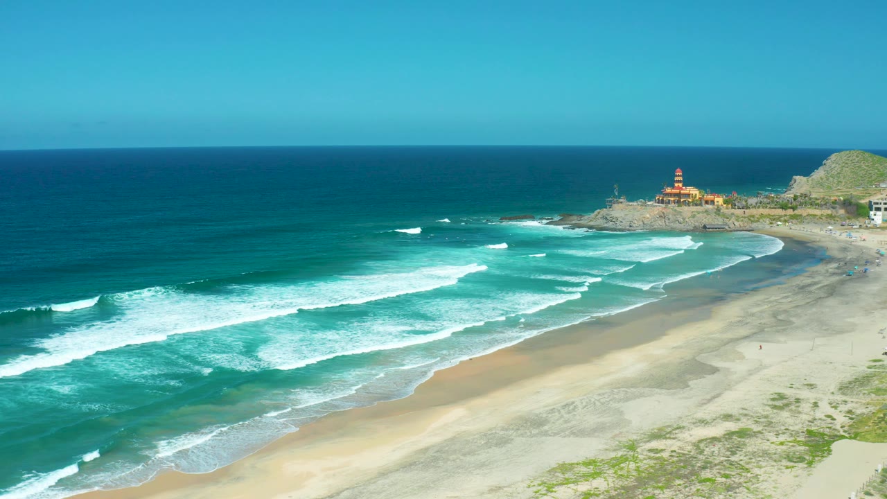 The beach and surf zone fill the frame as whitecaps break diagonally across Cerritos Beach, with the lighthouse and rocky point visible under clear sky and no visible people.