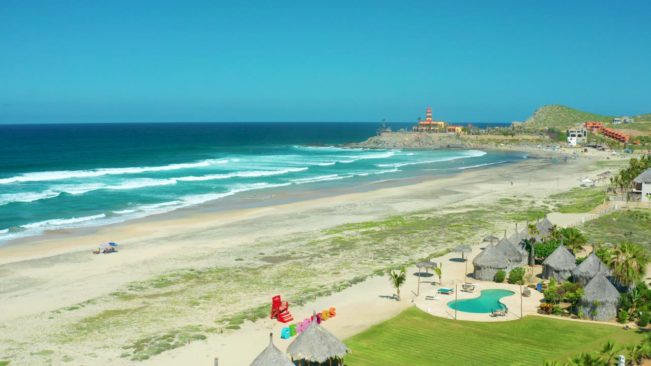 Cerritos Beach in Baja California Sur with turquoise waves, pale sand, green dune patches, a red and white lifeguard umbrella, a pool, thatched huts, and no visible people.