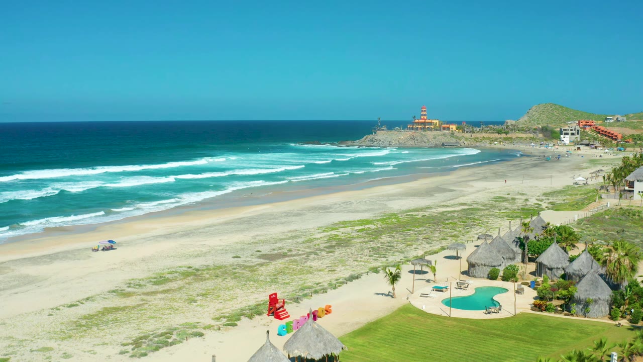 Sandy shoreline with rolling waves, pool, huts, and lifeguard umbrella.