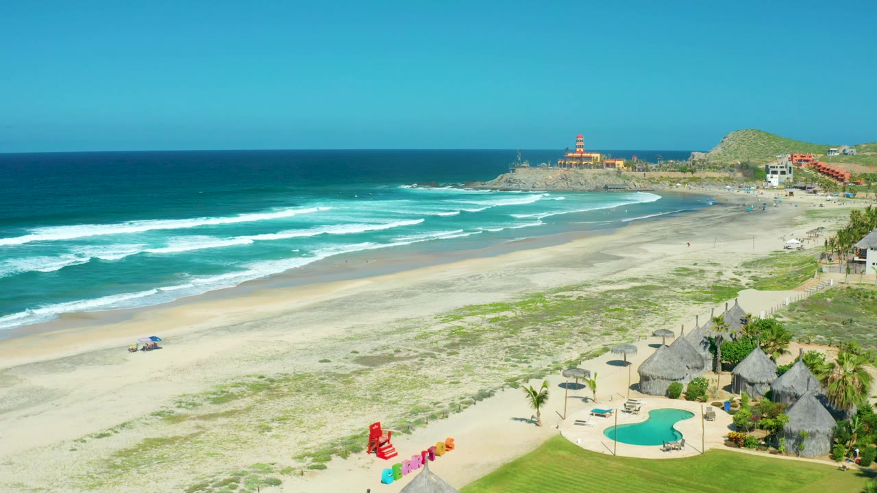 Cerritos Beach aerial frame with turquoise ocean on the left, white wave sets across the sand, a pool and thatched huts at the lower right, distant buildings, a rocky point, and no visible people.