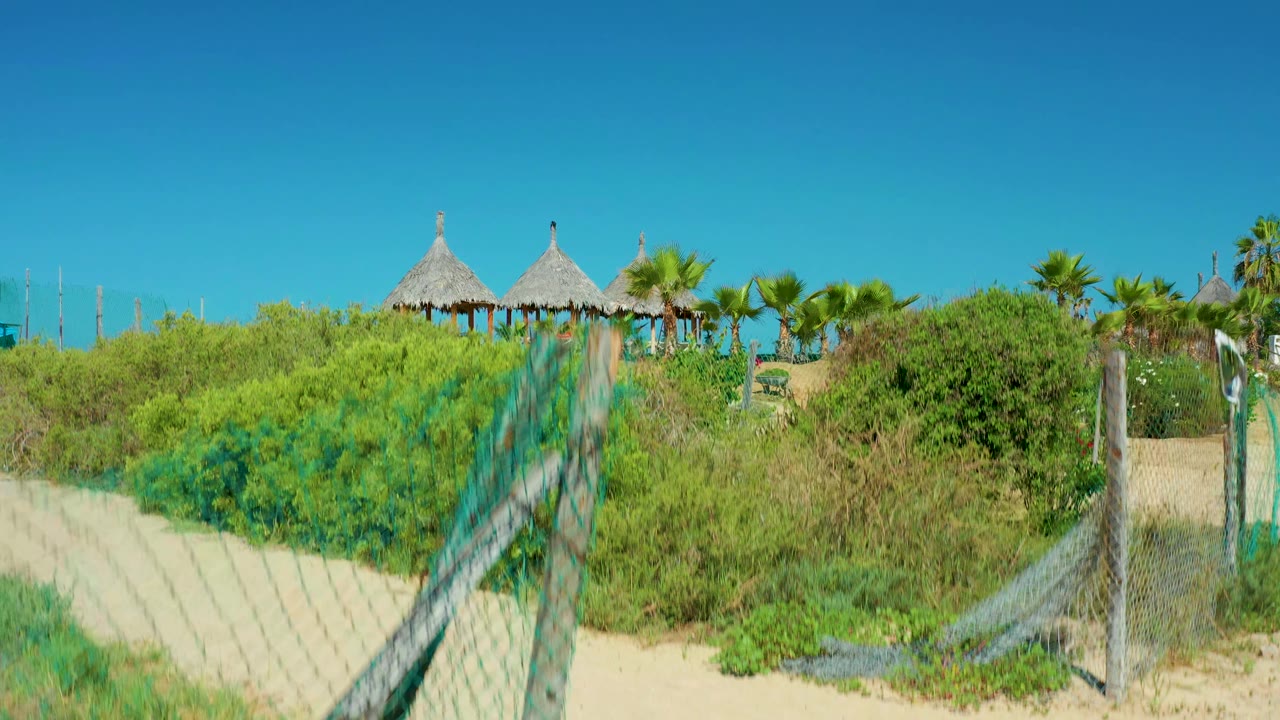 Cerritos Beach view through a chain-link fence, with pale sand, thick green vegetation, thatched hut roofs, palms, and no visible people.