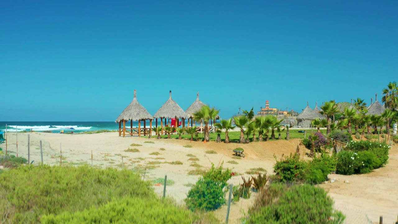 Cerritos Beach opens to pale sand, ocean surf, thatched huts on posts, palm plantings, resort buildings in the distance, and no visible people.