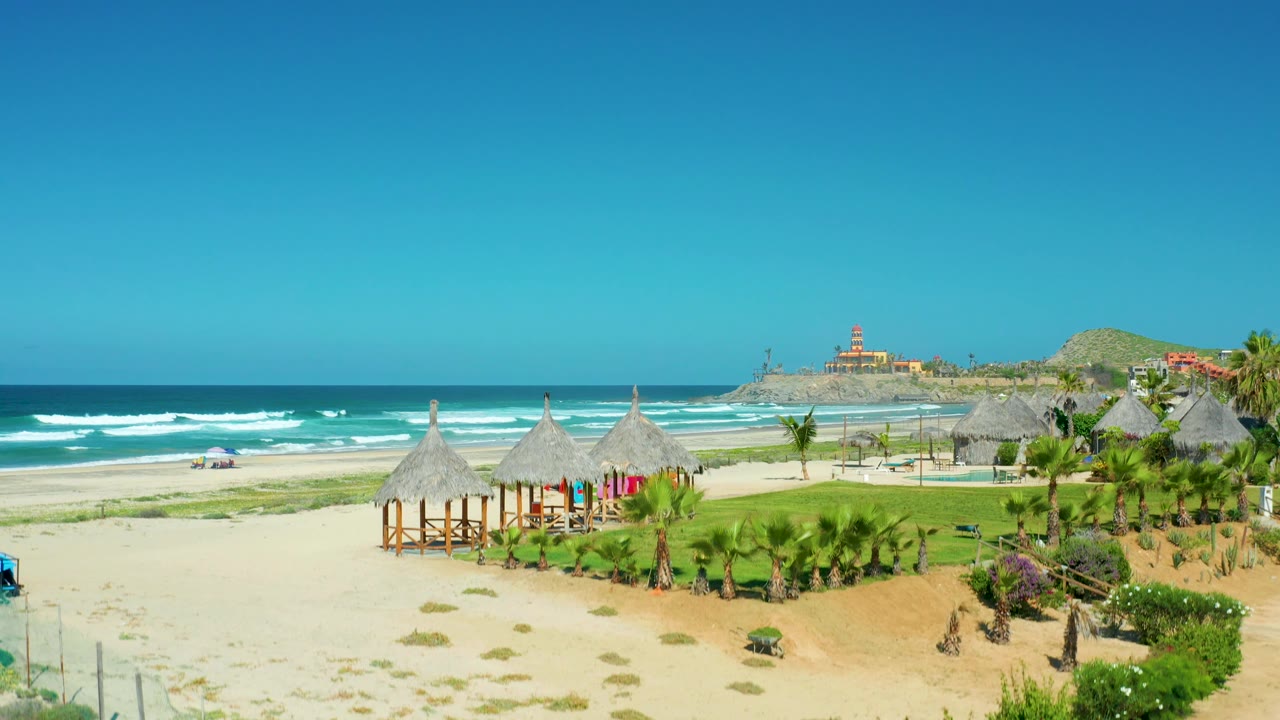 Wide beach and ocean view with cabanas, palms, and distant hill.