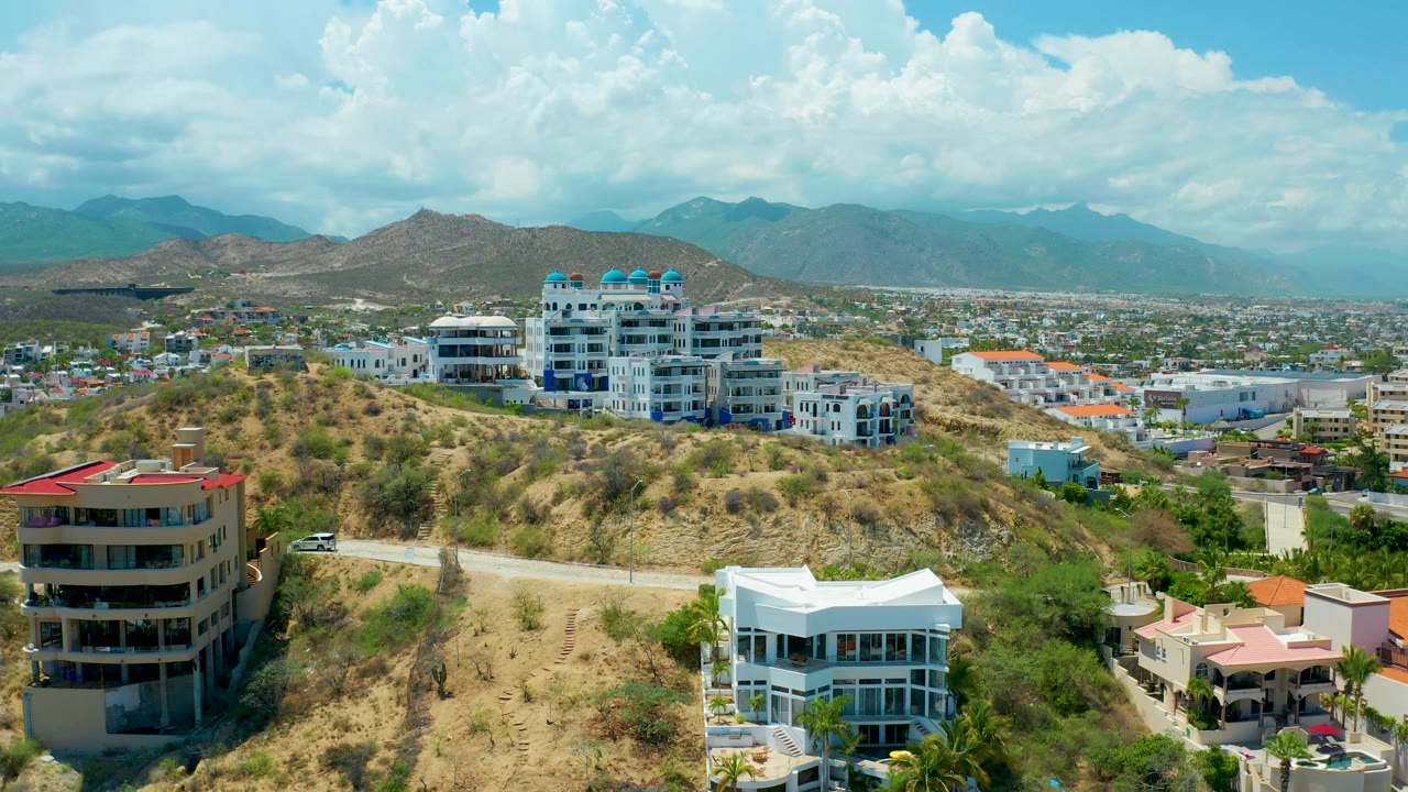 Wide elevated view of a San Jose del Cabo hillside neighborhood with modern residential buildings, a winding road, distant city blocks, blue mountains, large clouds, and no visible people.