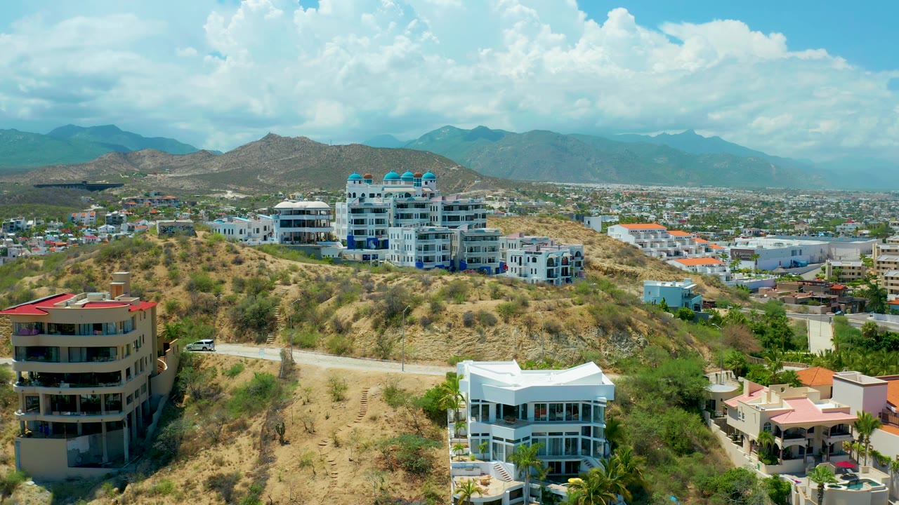 Elevated San Jose del Cabo view with the white hillside residence lower right, a darker apartment building lower left, a pale road across the slope, distant city blocks, mountains, clouds, and no visible people.