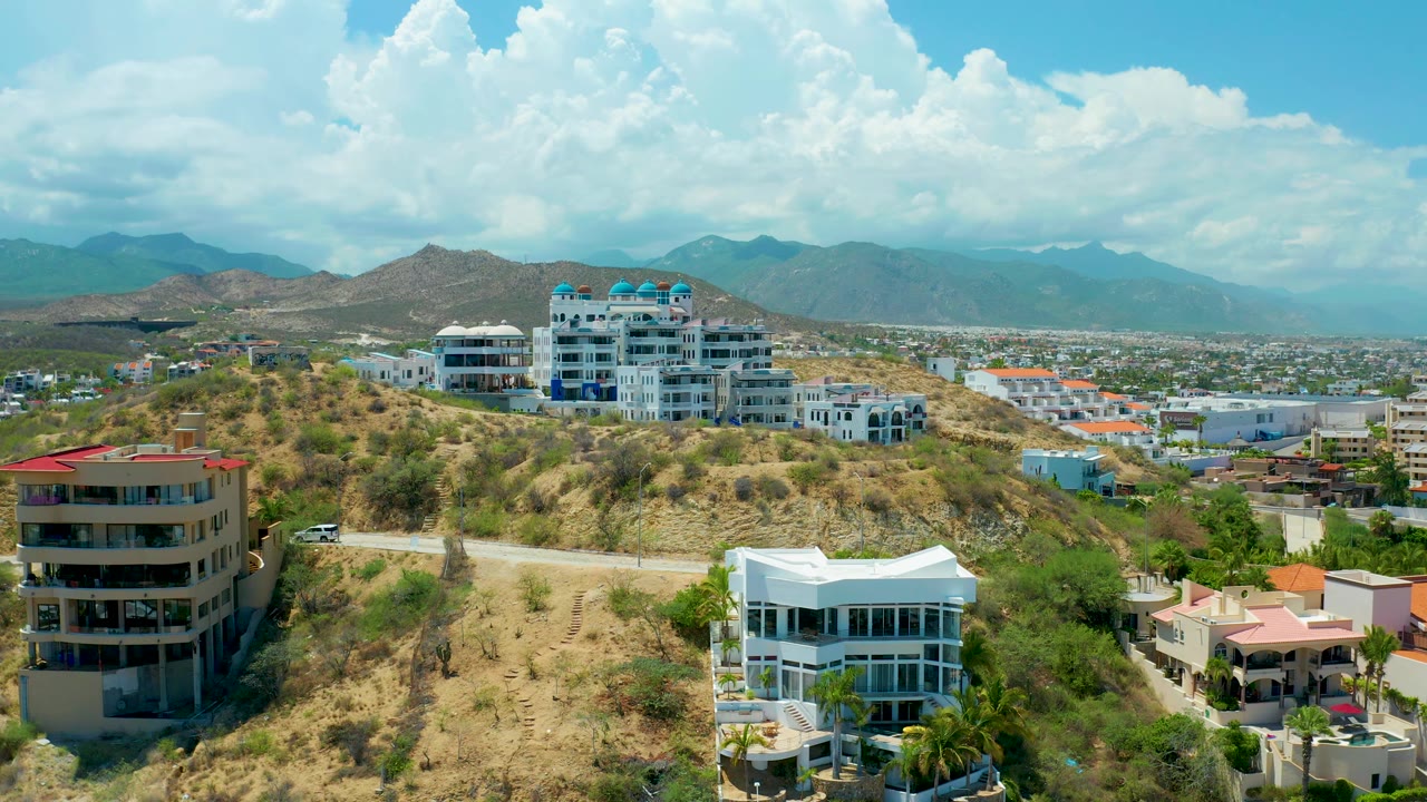 Residential hillside with a road, white house, darker building, distant city, and mountains.