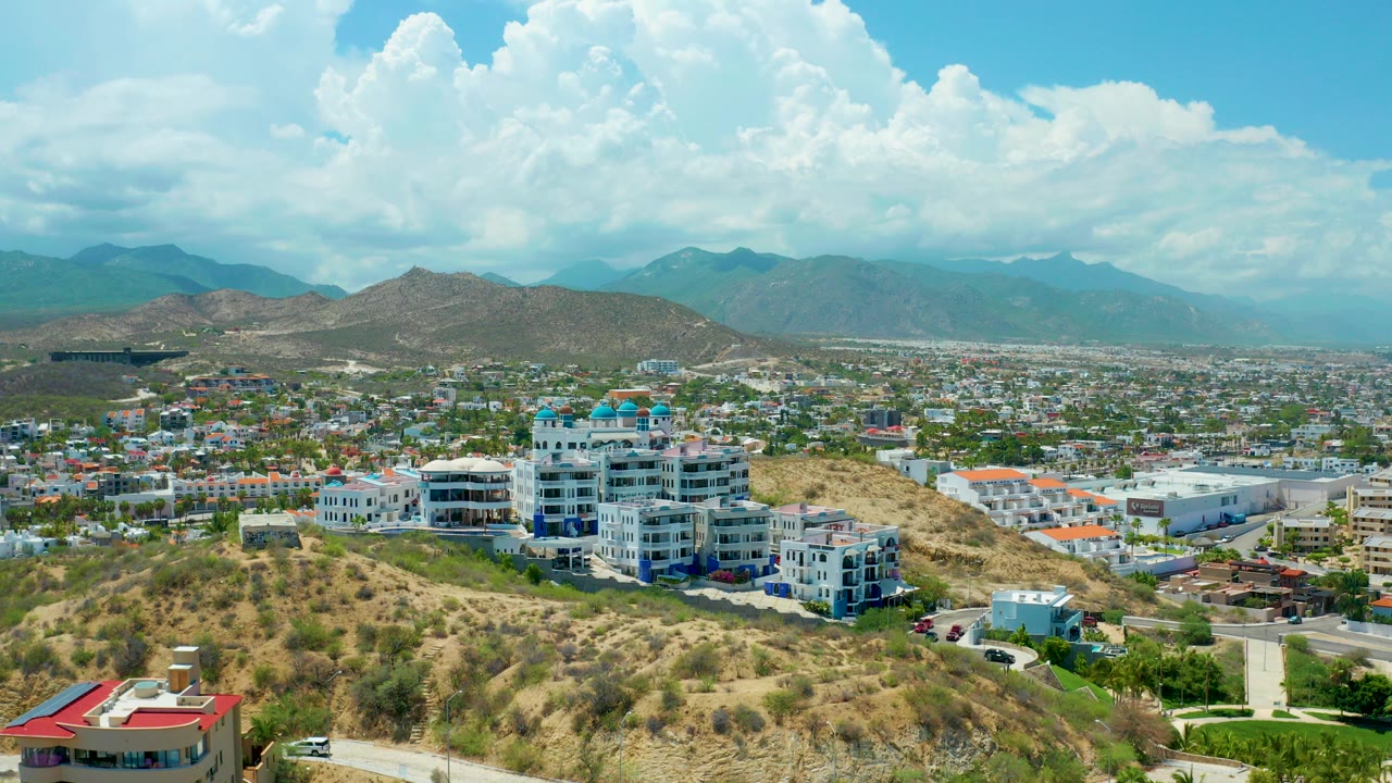 Opening aerial frame over San Jose Del Cabo, Mexico, with dry hillside brush, white and blue buildings on the slope, city blocks beyond, mountains in the distance, and no visible people.