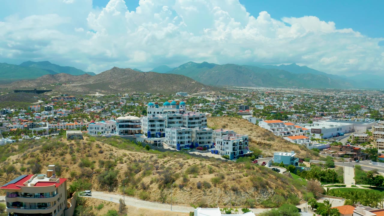 Aerial view over San Jose Del Cabo with clustered white hillside buildings at mid-frame, tan scrub-covered slopes in front, town streets behind, hazy mountains, and no visible people.