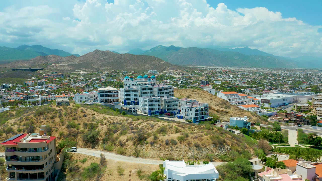 Aerial city edge view with hillside buildings, dry vegetation, and clouded mountains