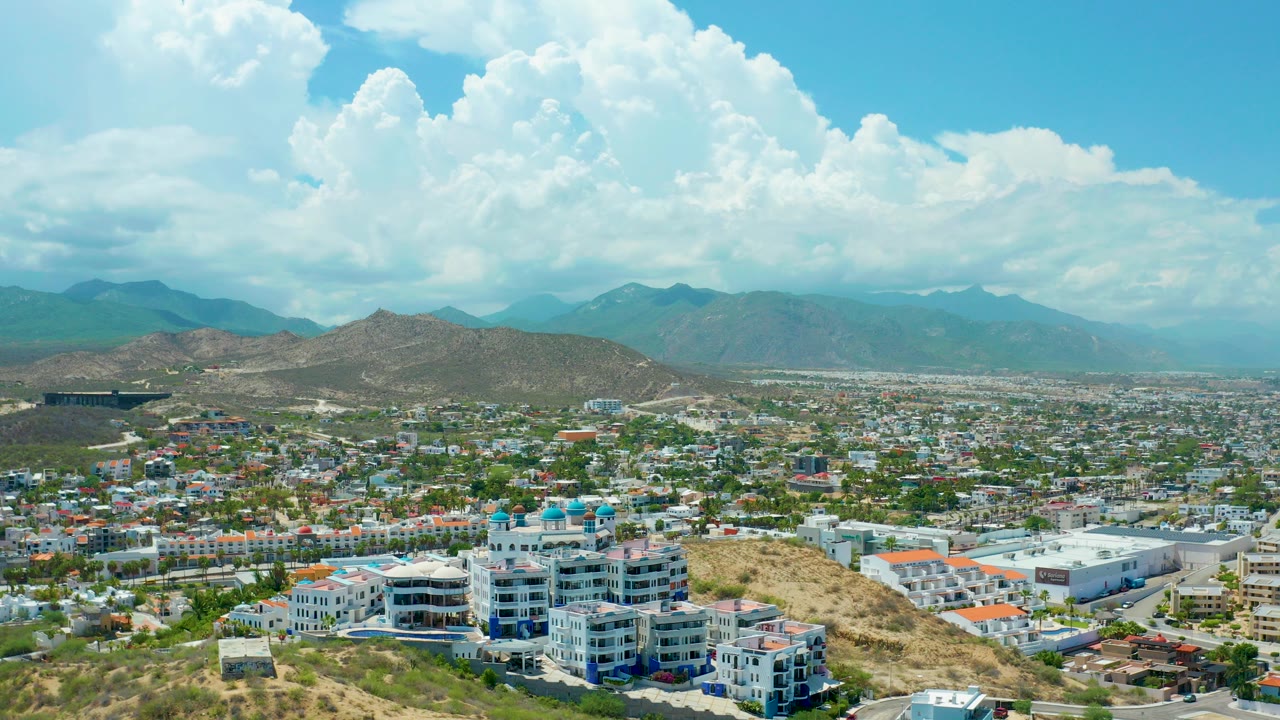 Opening frame of San Jose del Cabo showing hillside buildings, dry terrain, a broad city grid, distant mountains, large white clouds, and no visible people.