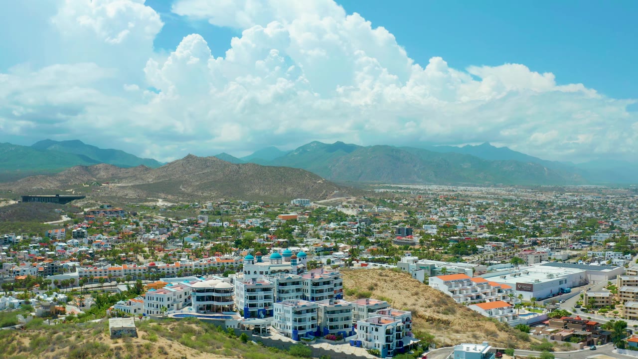 San Jose del Cabo aerial frame with white foreground buildings, dry hillside brush, low city blocks, hazy mountains, broad cumulus clouds, and no visible people.