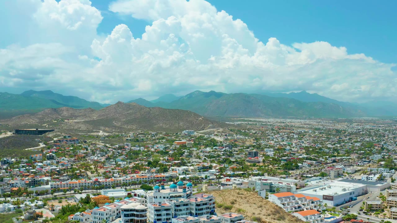 Aerial view over San Jose del Cabo with hillside buildings in the foreground, a broad city grid, distant mountains, large white clouds, and no visible people.