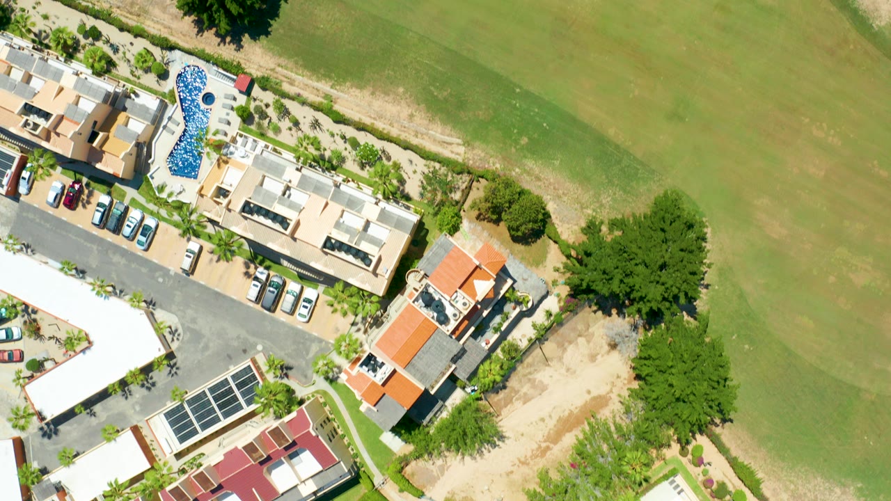 Top-down aerial frame in San Jose Del Cabo showing white villas, a blue pool, parked cars, solar panels, and golf course grass with no visible people.