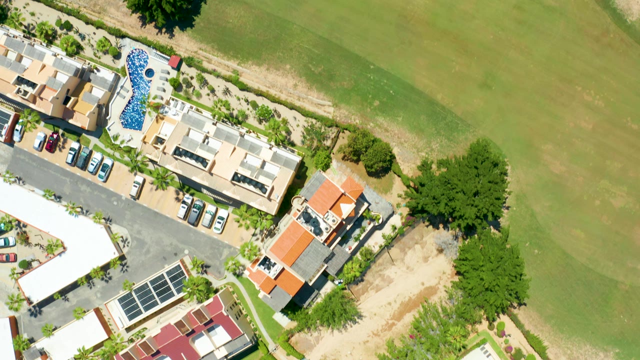 High vertical aerial frame in San Jose Del Cabo with a long blue pool, clustered villas, parked cars, trees, and open green golf course area, with no visible people.
