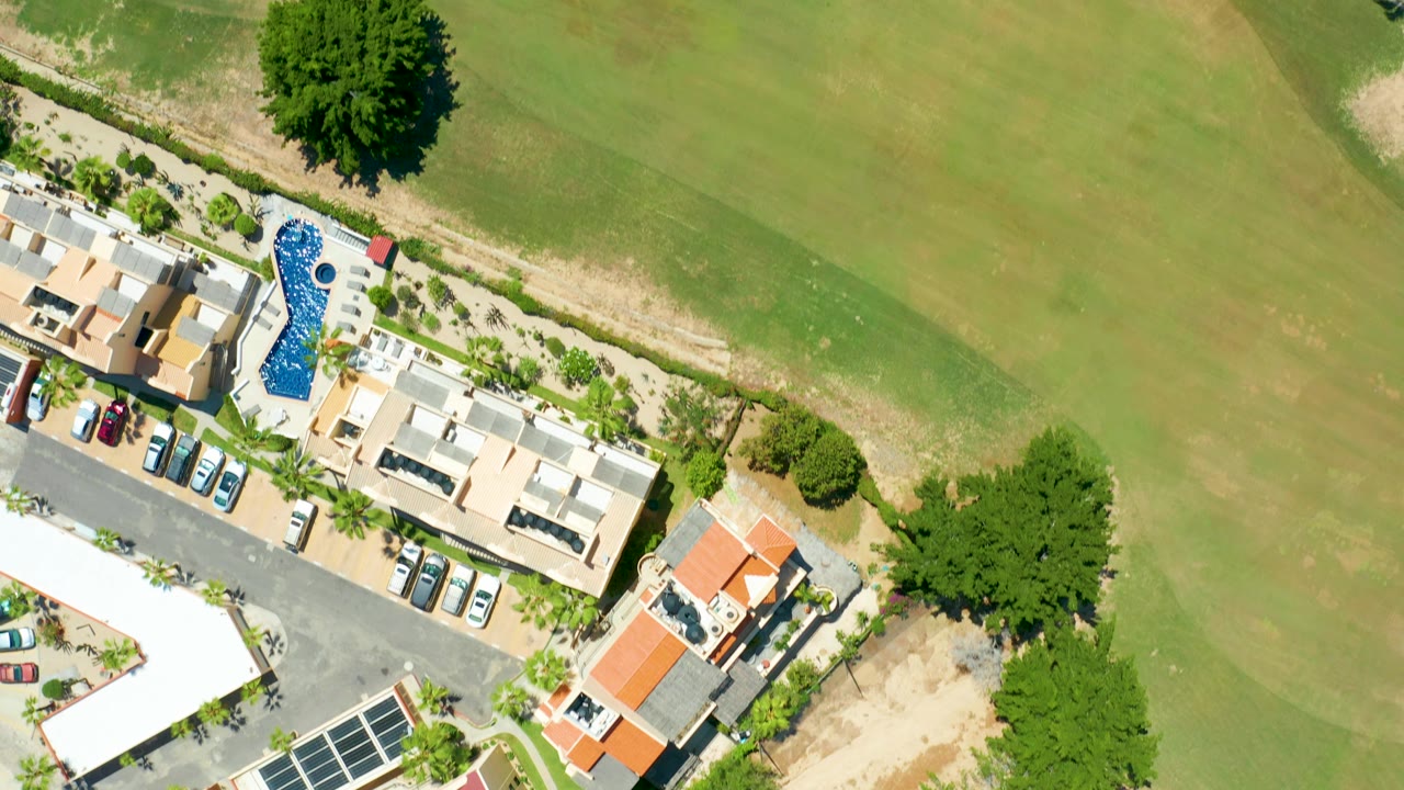 Aerial frame in San Jose Del Cabo showing residential rooftops and parking at left, a blue pool near landscaped paths, and broad golf course grass to the right, with no visible people.