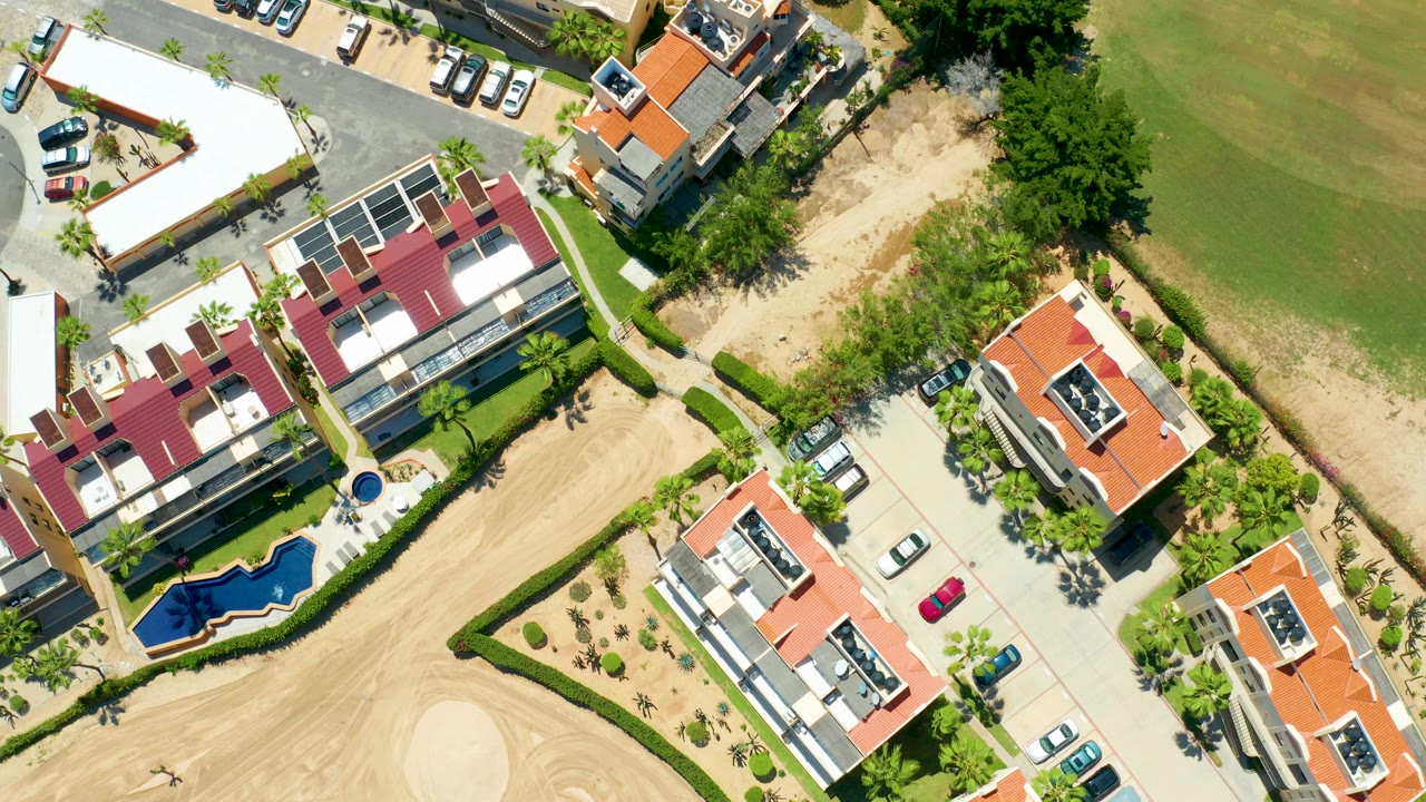 Top-down aerial frame from San Jose del Cabo showing red-roof and white-roof homes, parked cars, a small blue pool, sandy ground, trees, and a green field, with no visible people.
