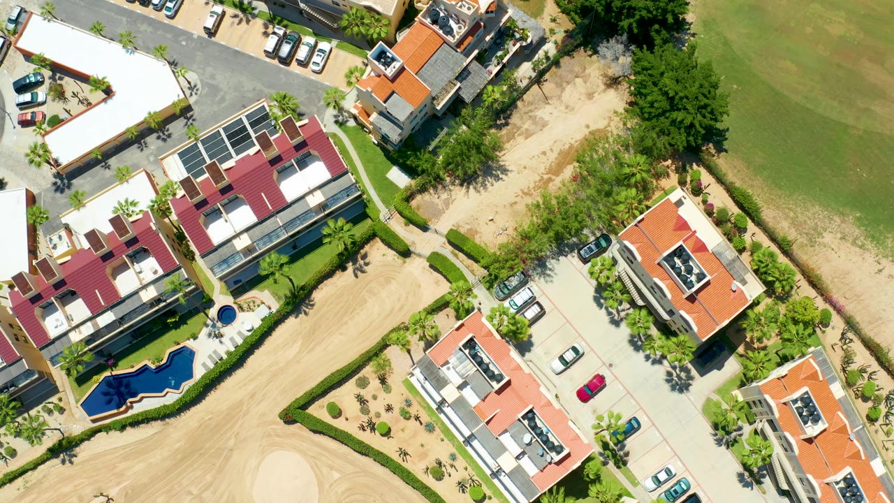 At 0:02, the San Jose del Cabo drone frame shows a similar residential cluster shifted across the frame, with red and orange roofs, parked cars, trees, sandy strips, a blue pool, and no visible people.