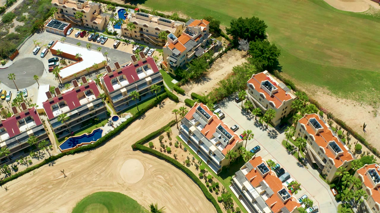 Drone view over residential buildings, parking, pool, sand, and fairway grass.