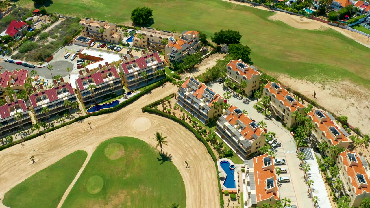 High aerial view in San Jose del Cabo showing red-roof residential buildings, blue pools, parked cars, palms, green fairway grass, and pale sand bunkers near the start of the clip.