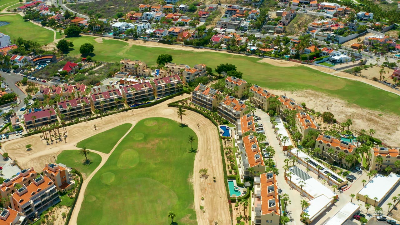 Aerial frame over San Jose Del Cabo showing a green golf fairway, sandy paths, red-roof homes, streets, small pools, and no visible people.