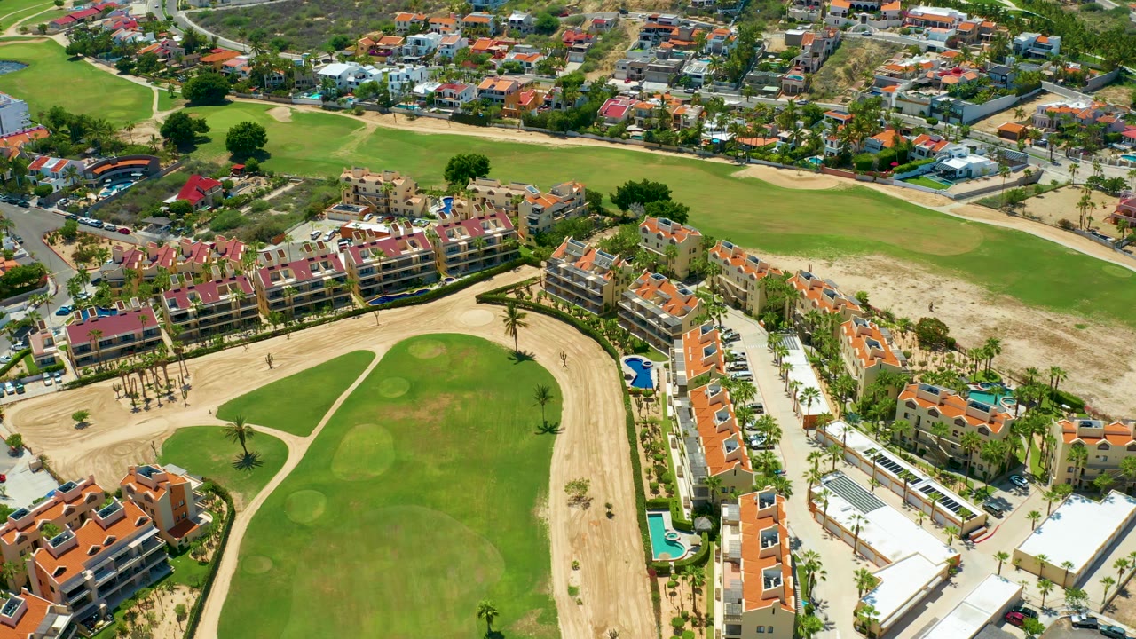 Aerial frame in San Jose Del Cabo with the fairway shifted across the lower view, tan sandy areas, red and orange roof housing, pools, and no visible people.