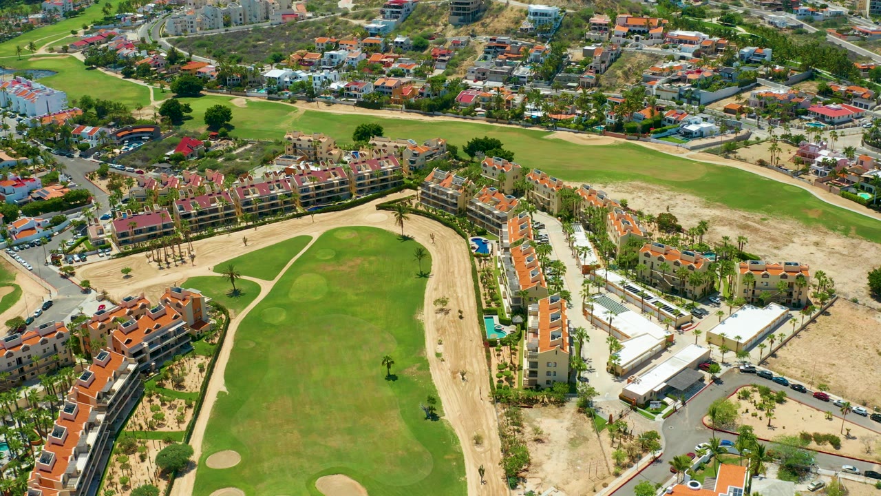Aerial frame over a San Jose Del Cabo golf course with a green fairway, sand maintenance areas, tiled-roof residences, roads, parked cars, palm trees, and no visible people.