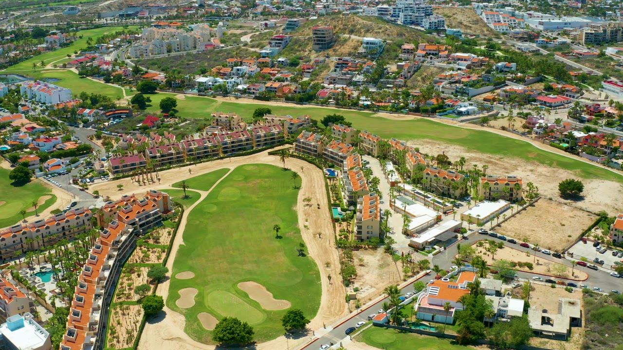 Aerial frame in San Jose del Cabo showing more of the fairway center, pale bunkers, roadside buildings, red-tile roofs, dry ground, and no visible people.
