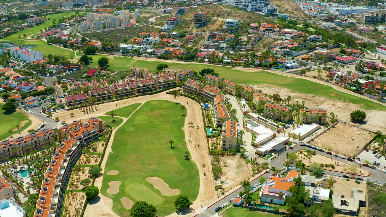 Rotating aerial view over San Jose del Cabo with the golf green shifting slightly in frame, tan undeveloped ground along the right, homes beyond, and no visible people.