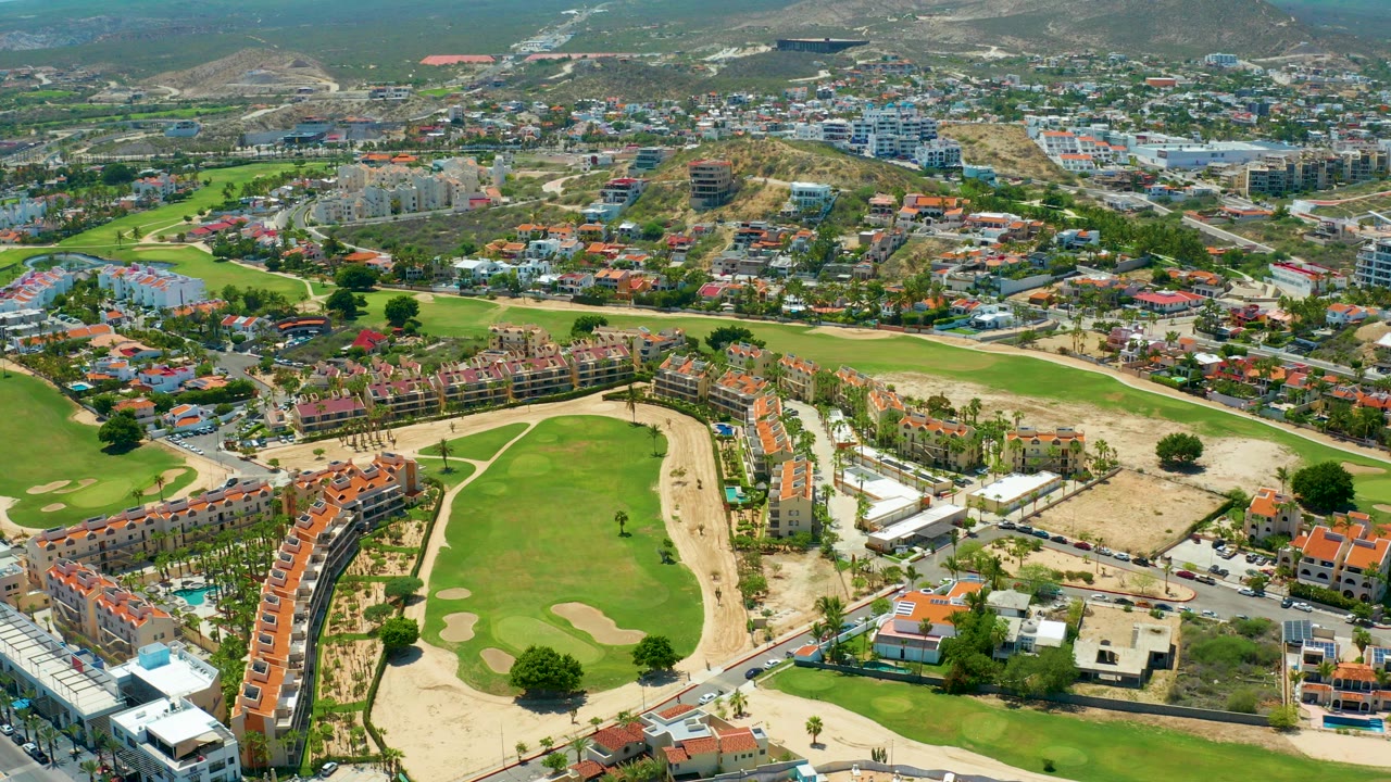 Aerial view in San Jose del Cabo, Mexico showing a bright golf green, sand bunkers, curved red-roof residences, roads, and hillside buildings, with a tiny person visible on the green.