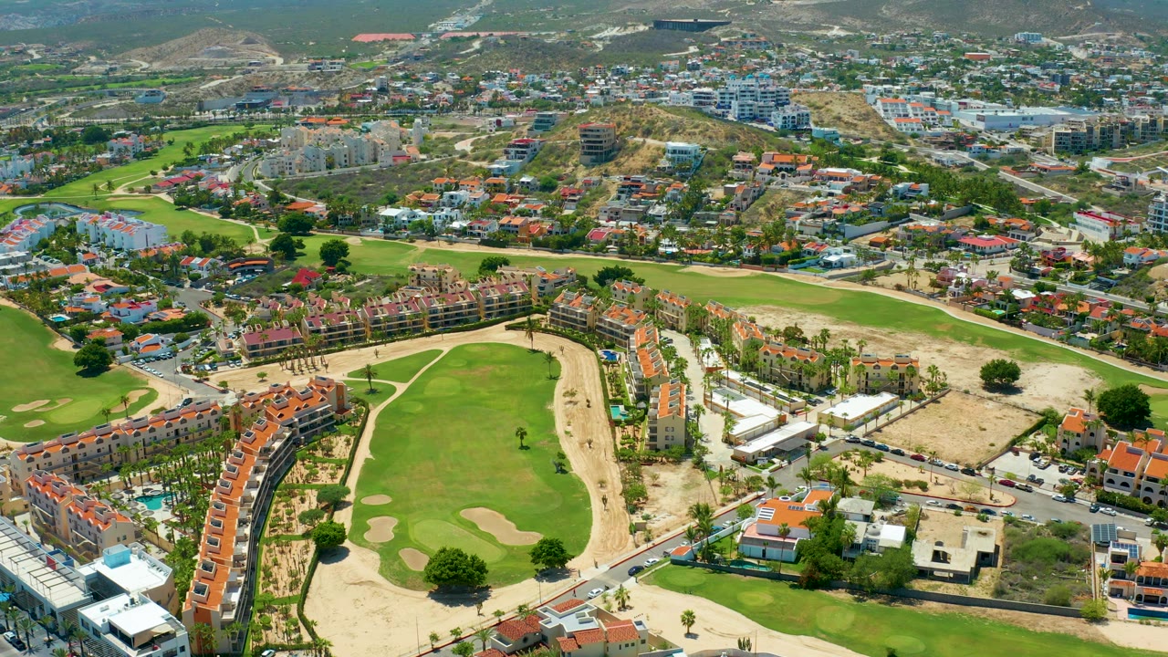 High aerial frame in San Jose del Cabo, Mexico showing connected golf greens, pale sand areas, curving neighborhood roads, red-roof homes, and layered hillside construction in bright sun.