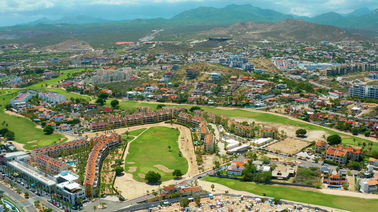 Aerial view over San Jose Del Cabo with a green golf course in the foreground, curved red-roof housing, roads, dry lots, distant hills, and no visible people.