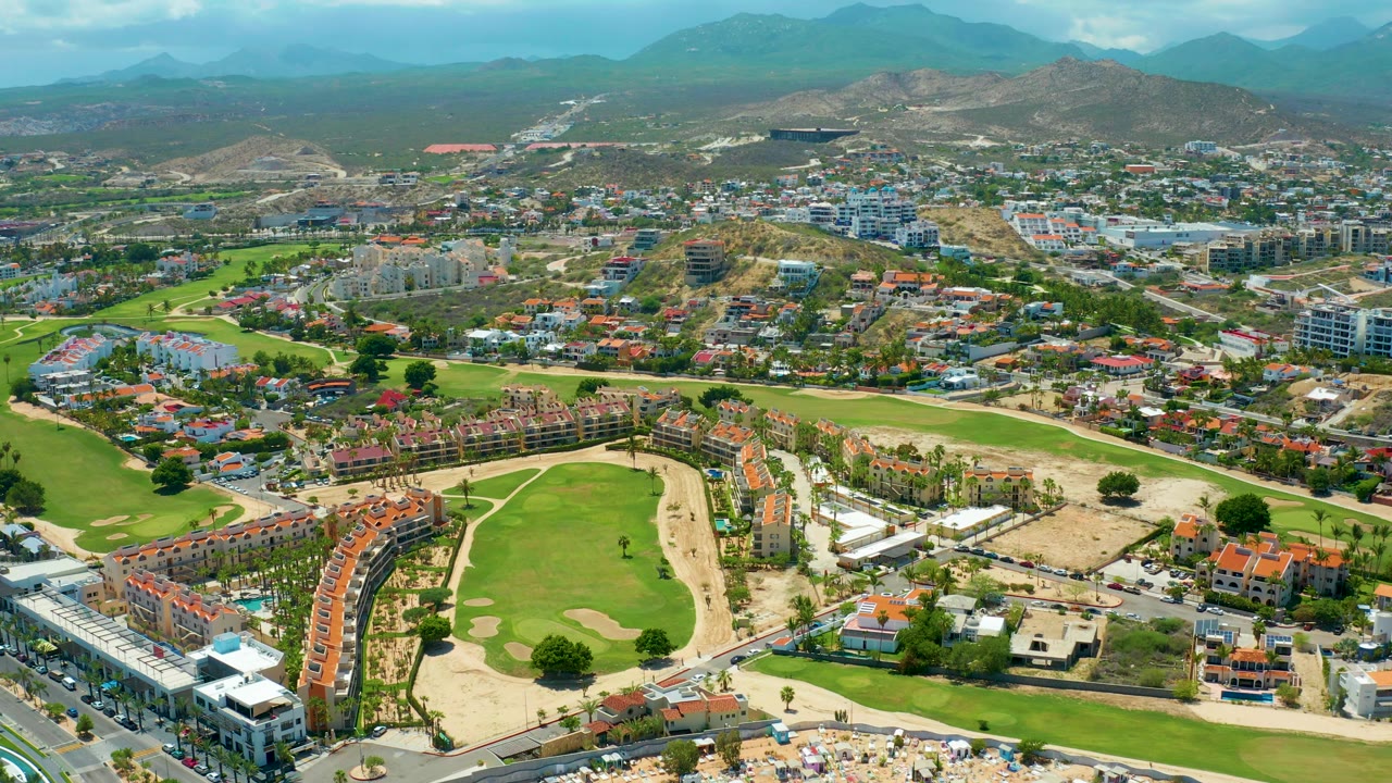 Aerial frame from San Jose Del Cabo with golf greens and sand areas in front, residential blocks across the middle distance, hazy hills, clouds, and no visible people.