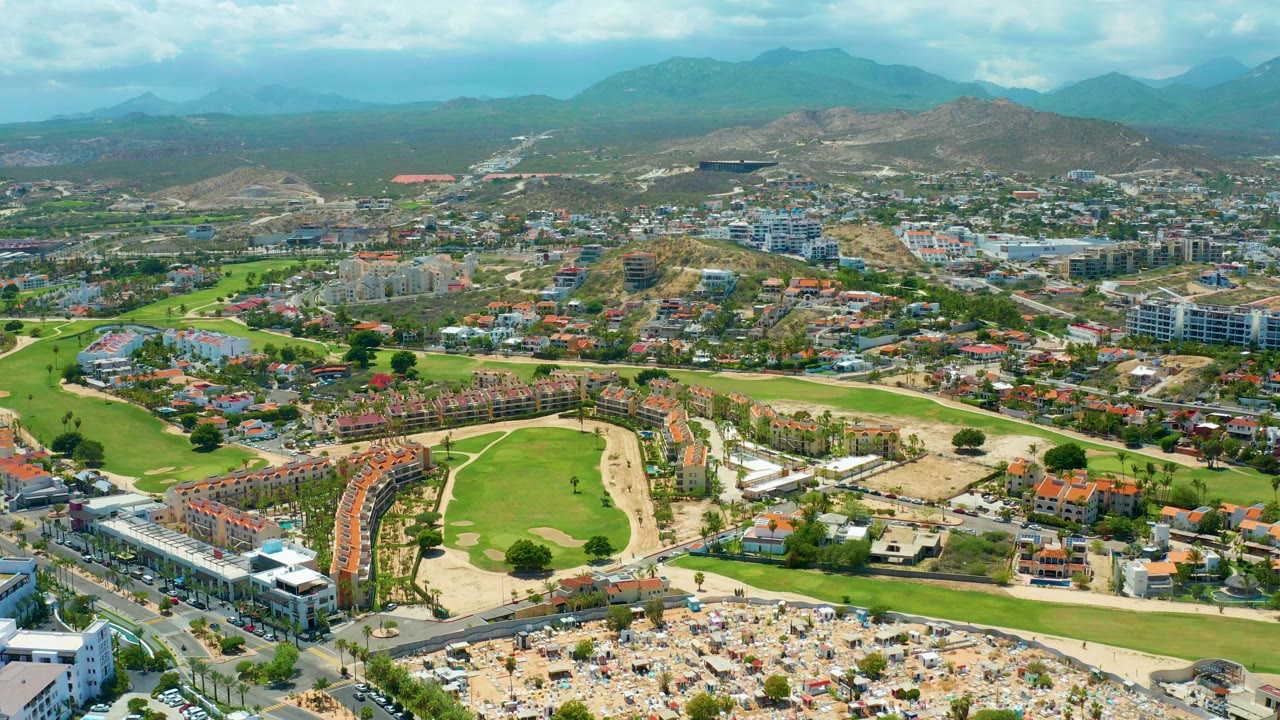 Aerial frame of San Jose del Cabo showing a green golf course, a sandy beach area with rows of umbrellas, resort buildings, roads, dry hillside terrain, distant mountains, and no visible people.