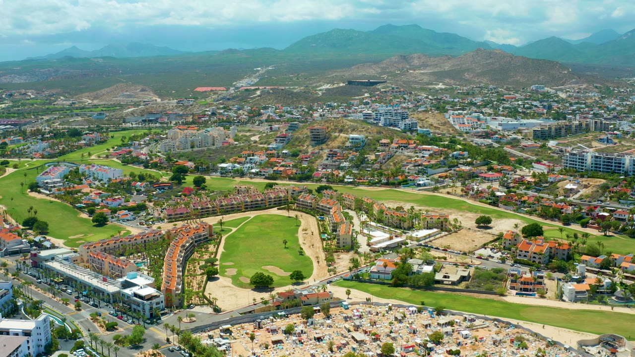 Wide drone frame over San Jose del Cabo with resort buildings and roads in the foreground, a golf course and sandy beach umbrella area across the lower half, urban blocks beyond, mountains on the horizon, and no visible people.