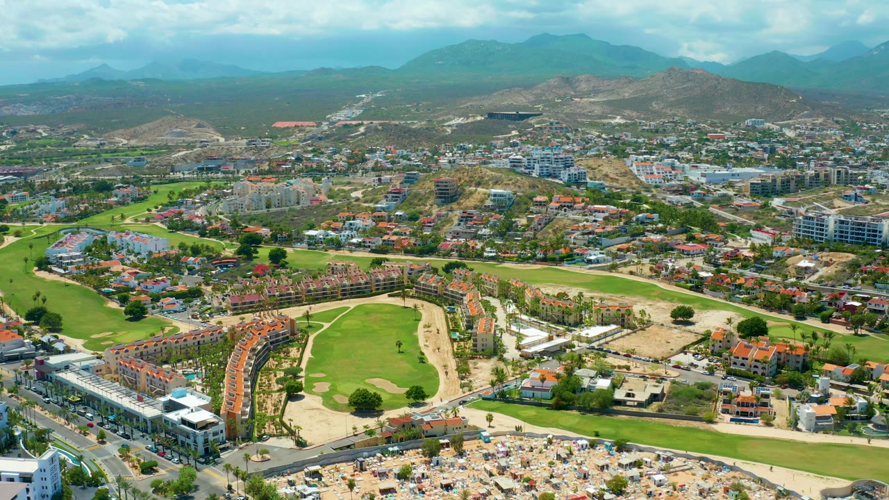Drone view of golf course beside resort buildings with beach umbrellas and mountains in the distance.
