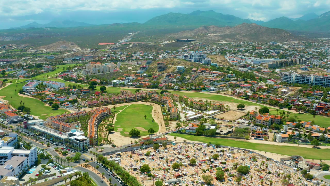 A high drone view of San Jose Del Cabo shows a curved golf course edge, nearby residential buildings, street grids, parking lots, mountains, and no visible people.